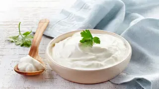 Un cuenco de cremosa crema agria decorado con perejil, junto a una cuchara de madera con más crema.