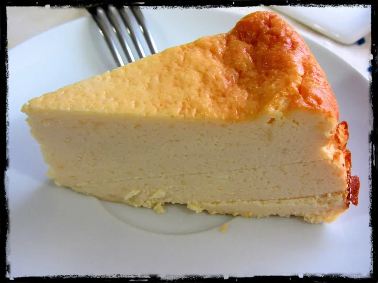 Una rebanada gruesa de tarta de queso cremosa, con la parte superior dorada y un tenedor al fondo.