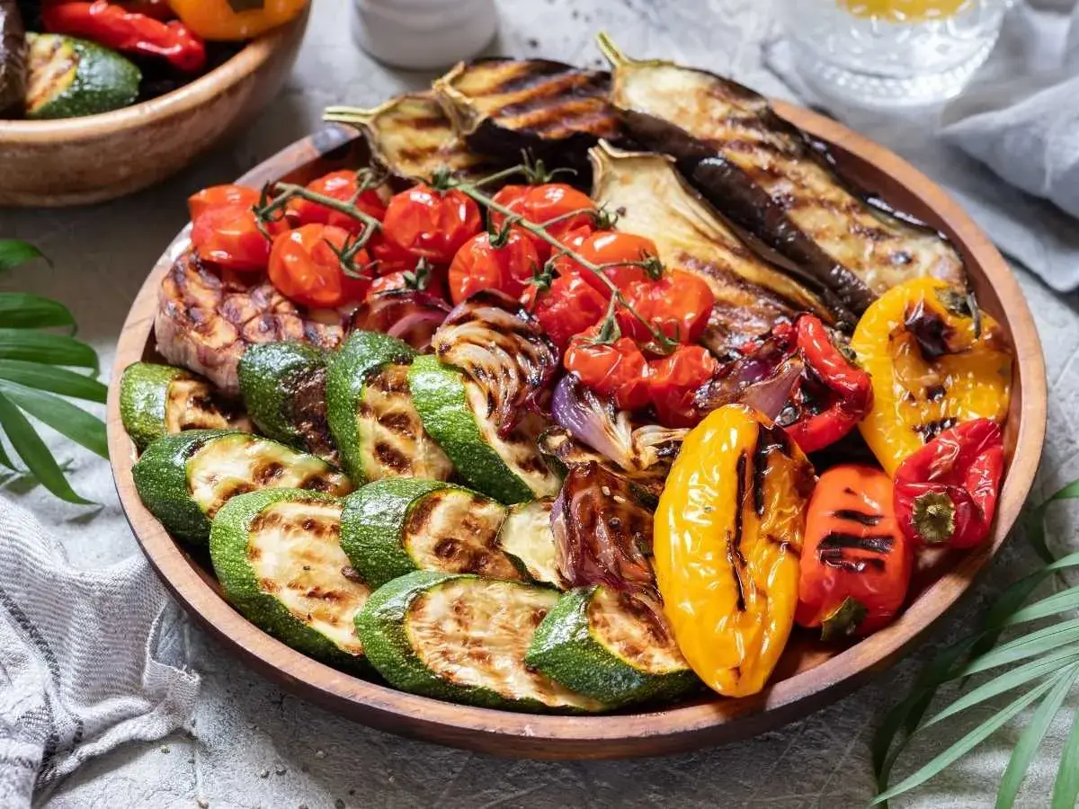 Una colorida parrillada de verduras con calabacín, berenjena, tomates cherry, pimientos y cebolla, servida en una bandeja de madera.