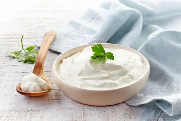 Un cuenco de cremosa crema agria decorado con perejil, junto a una cuchara de madera con más crema.