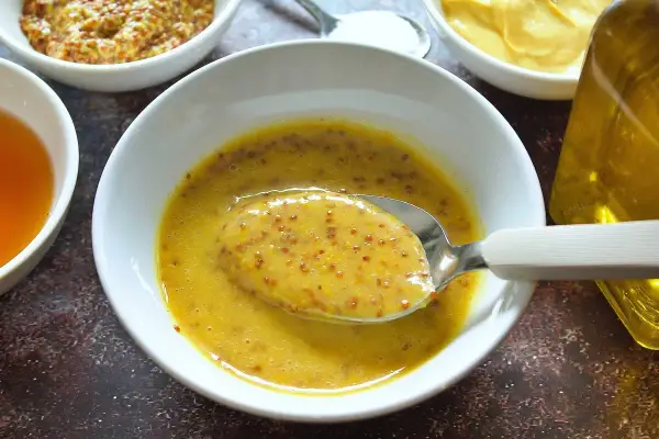 Cuchara con salsa miel y mostaza, lista para aderezar. Ingredientes en cuencos alrededor.