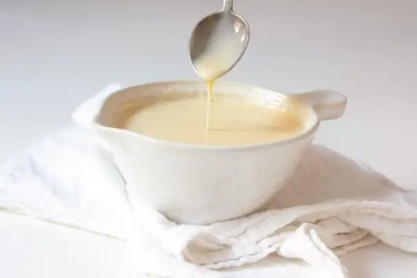 Cuchara vertiendo crema inglesa en un cuenco blanco sobre tela arrugada.