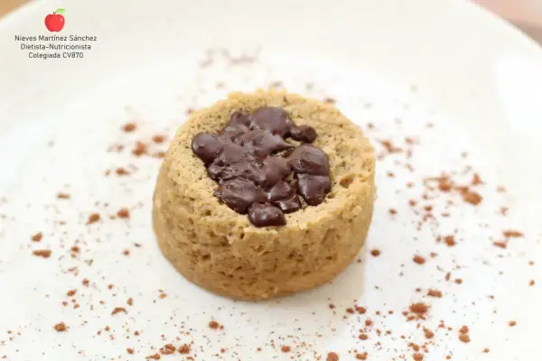 Delicioso mug cake saludable con trozos de chocolate negro, espolvoreado con cacao en polvo sobre un plato blanco.